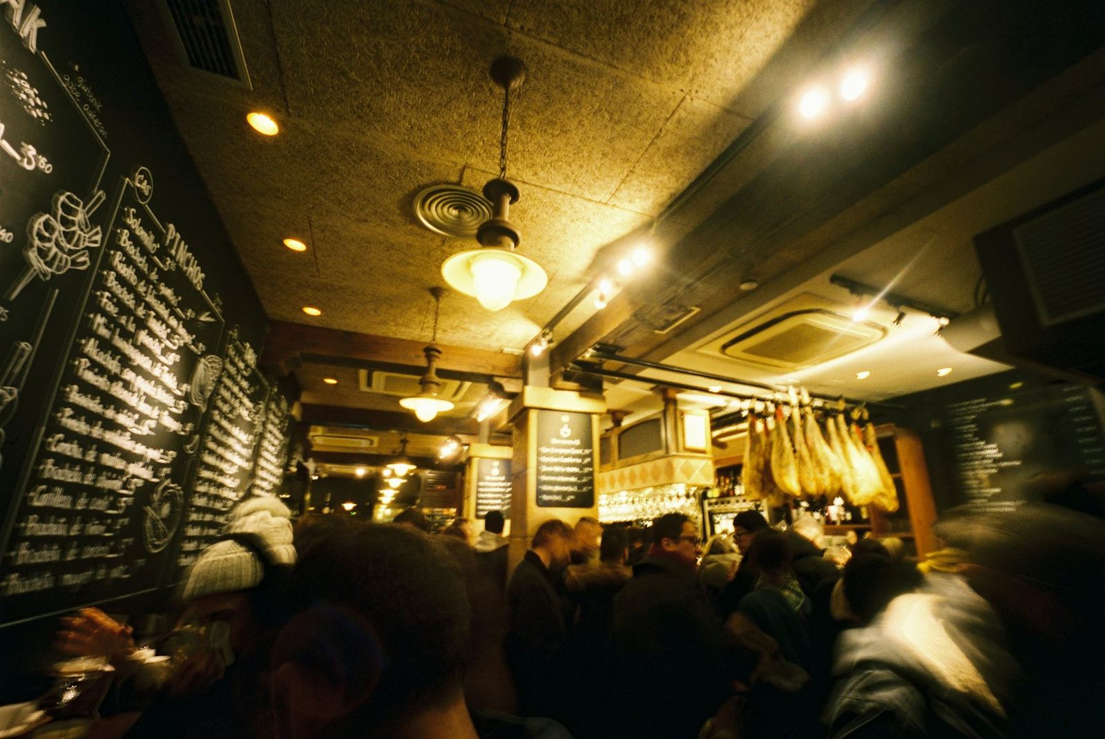 A busy tapas bar with hams hanging from the ceiling and boards written in chalk