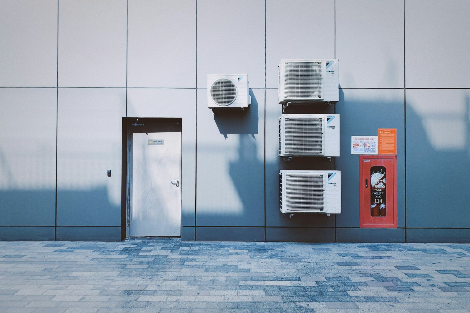 Grey façade with four outdoor units hung beside a door and a fire hose cabinet