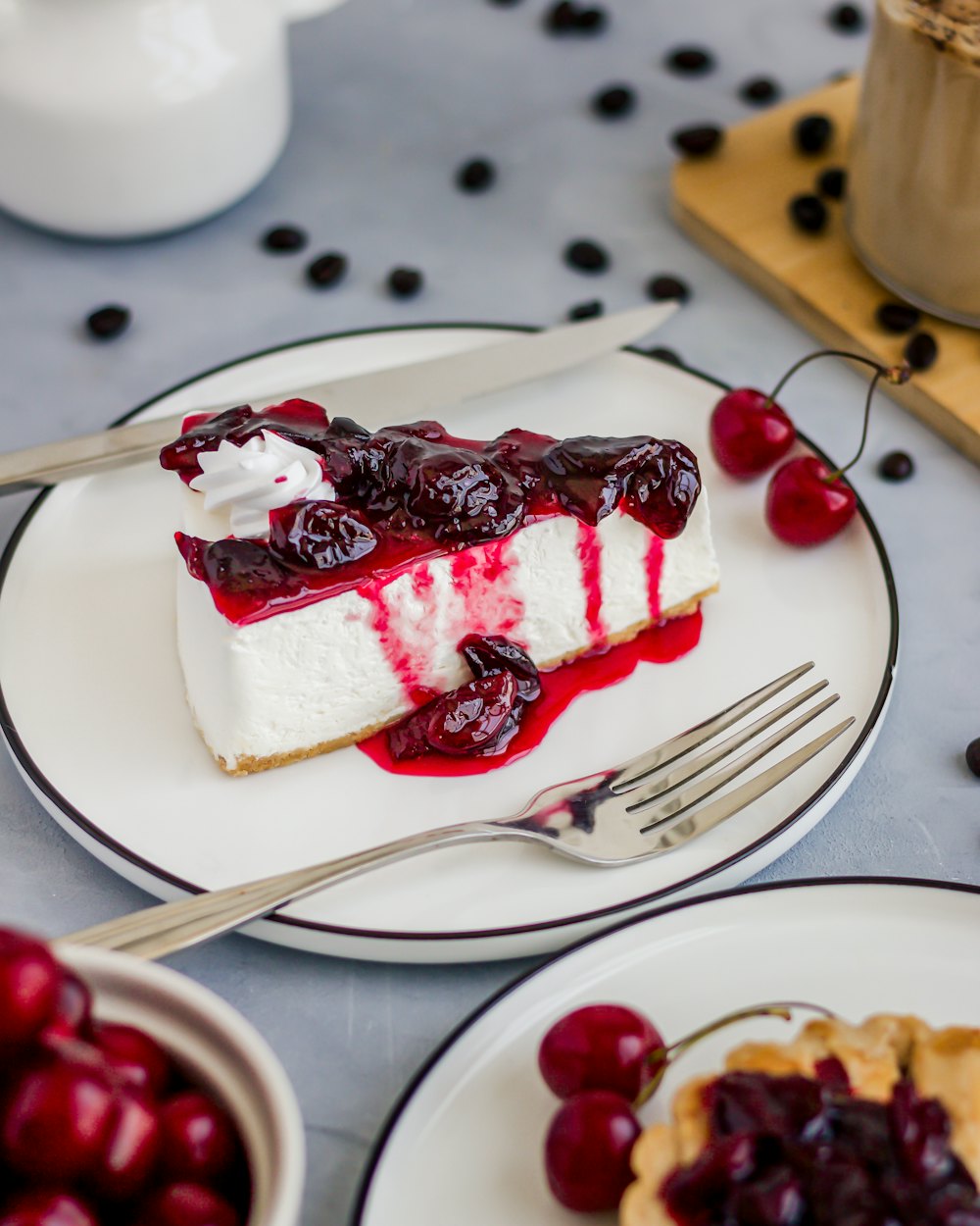 Porción de cheesecake con cerezas en almíbar, y cerezas frescas alrededor del plato